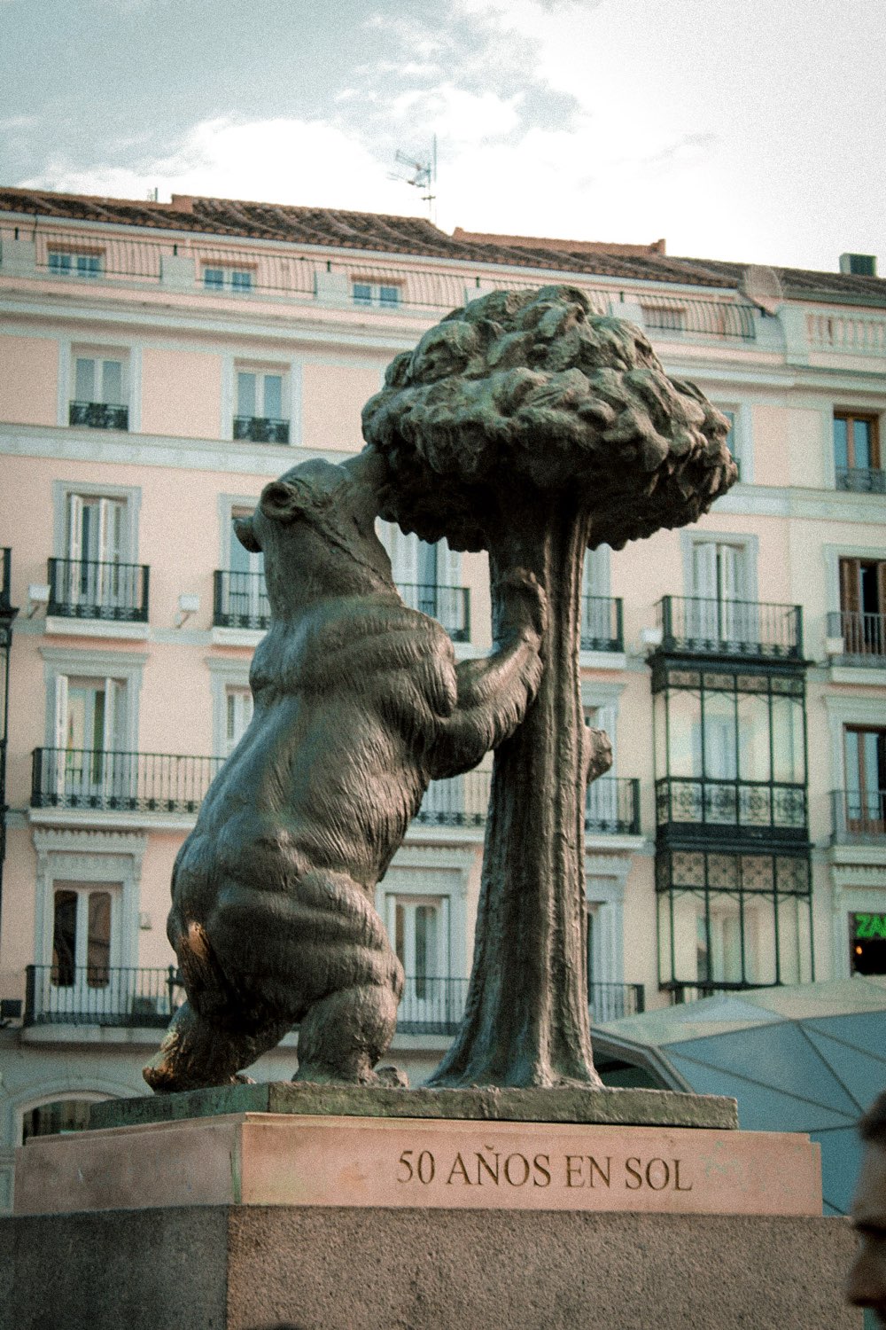 Bronze statue of a bear reaching up a strawberry tree, in front of a pale apartment façade with balconies