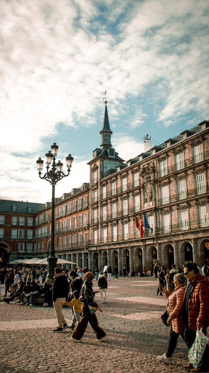 People crossing Plaza Mayor in Madrid past an ornate lamp post and arcaded buildings