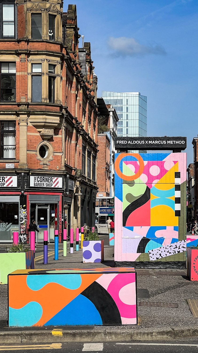 Brightly painted planters and a utility box on a Manchester street corner, red brick buildings behind