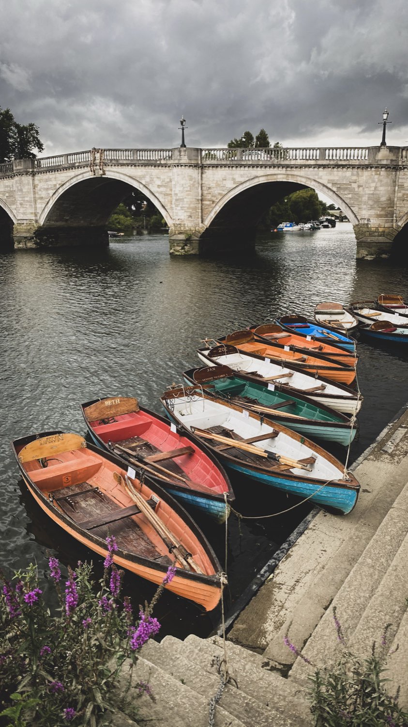 Wooden rowing boats moored in a row beside stone steps under an arched bridge on a grey day