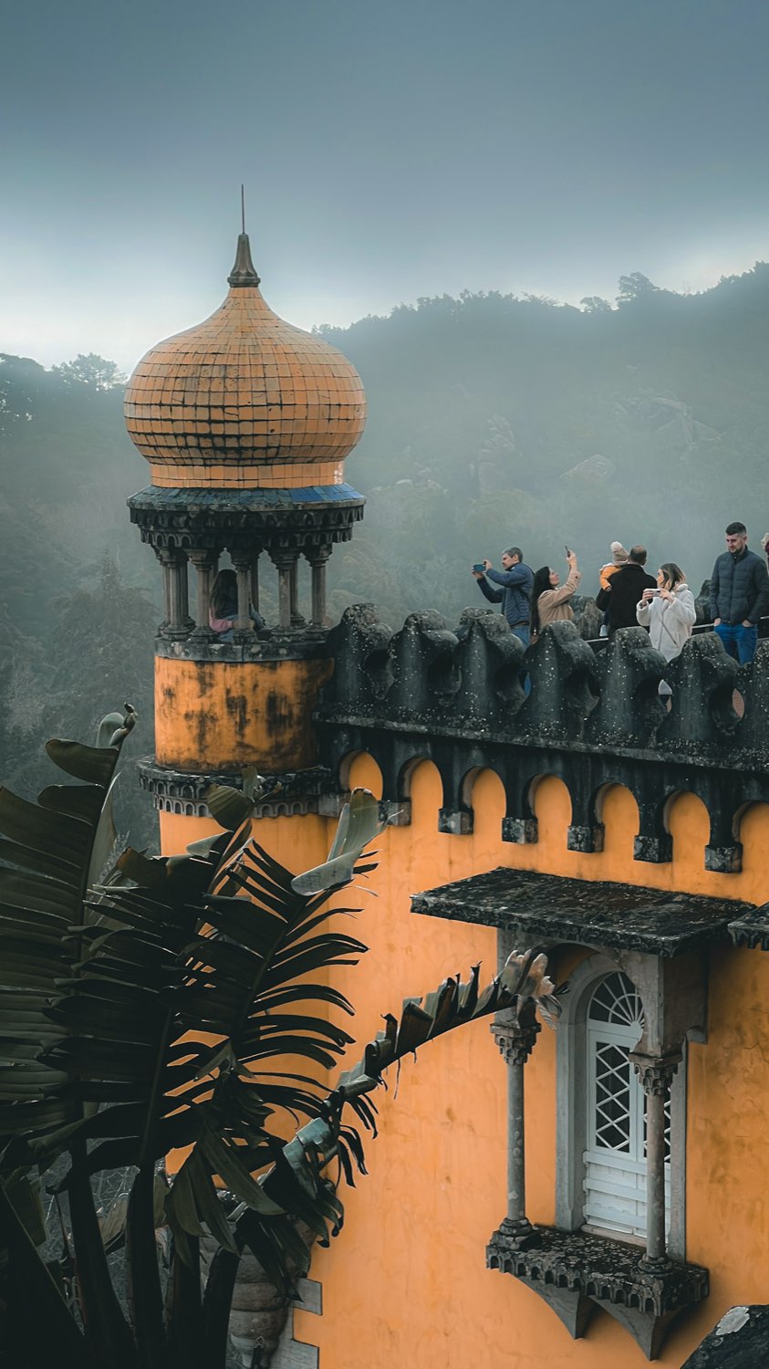 The ochre dome and battlements of the Pena Palace emerging from hill mist, with visitors on the terrace
