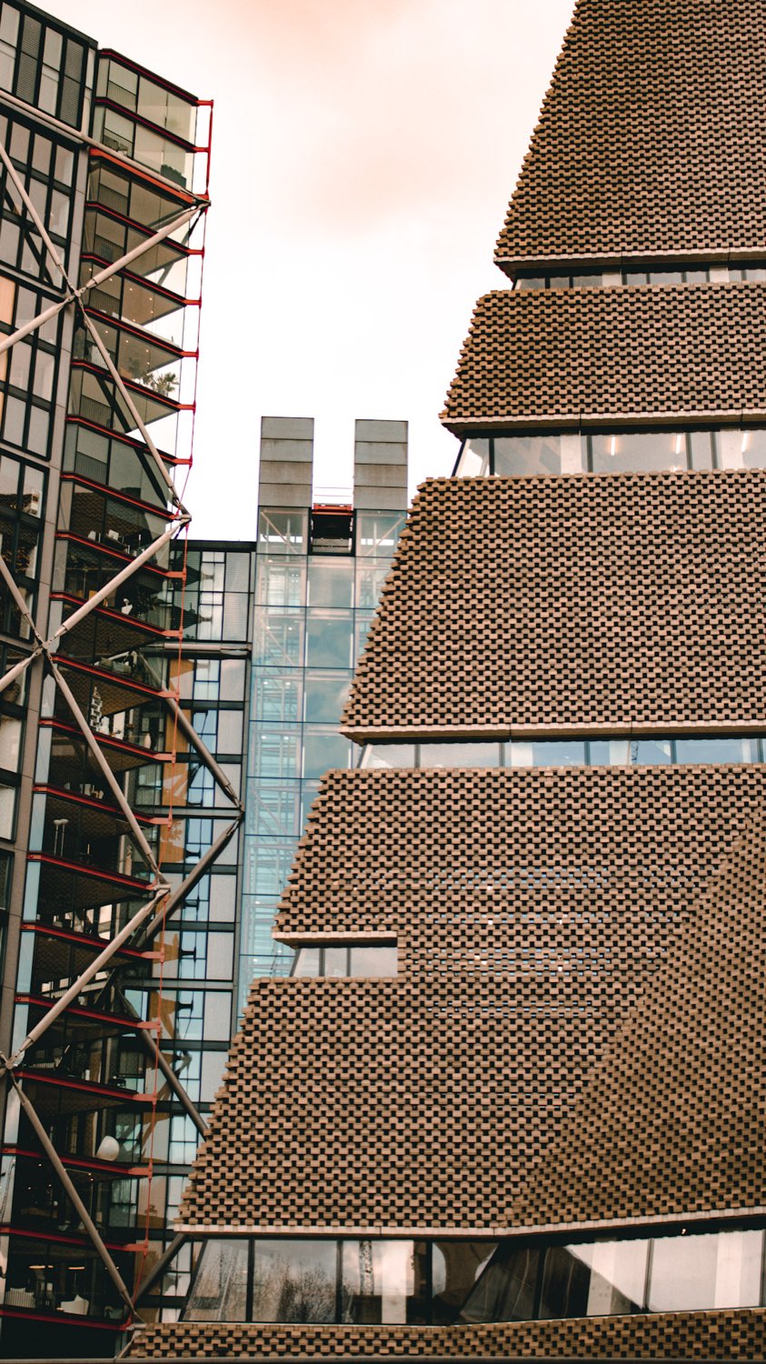 Angled perforated brick façade of the Tate Modern extension beside a glass building with red steel bracing