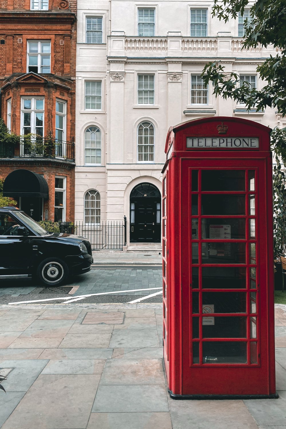 A red telephone box on a paved street beside a black cab and Georgian townhouses